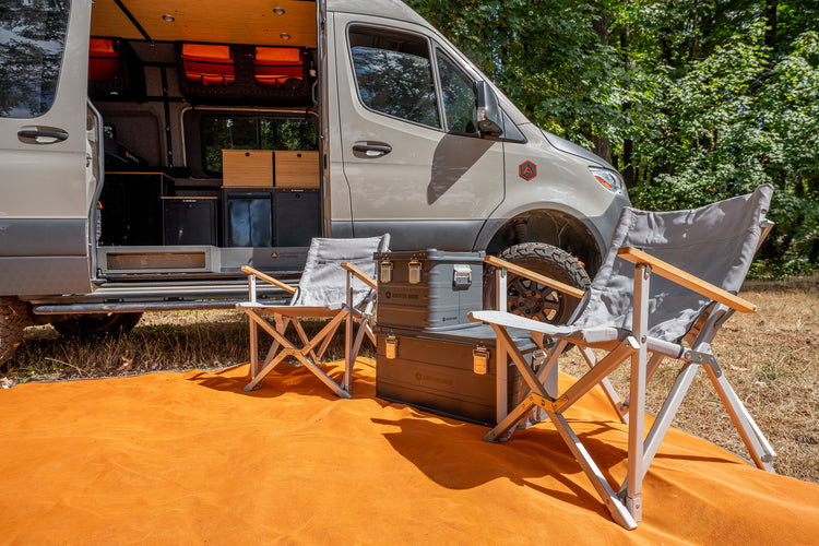 A gray camper van with its side door open, parked outdoors on a bright orange ground cover with two gray folding camp chairs and a metal storage trunk in front. Trees and grass are visible in the background.