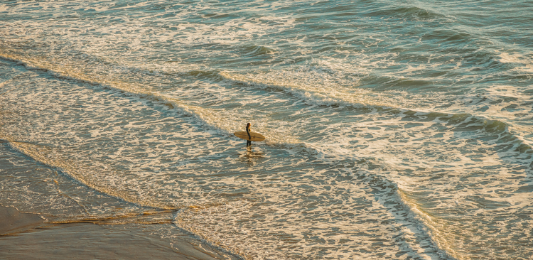 Surfer standing in waist-deep water at golden hour, looking out at the waves
