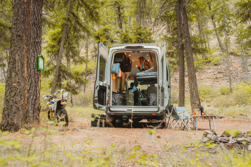Adventure Wagon Solo Nomad camper van parked at a forest campsite with rear doors open.