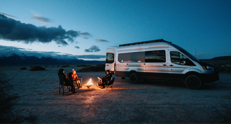 Three people sitting around a campfire with an Adventure Wagon sprinter van behind them 