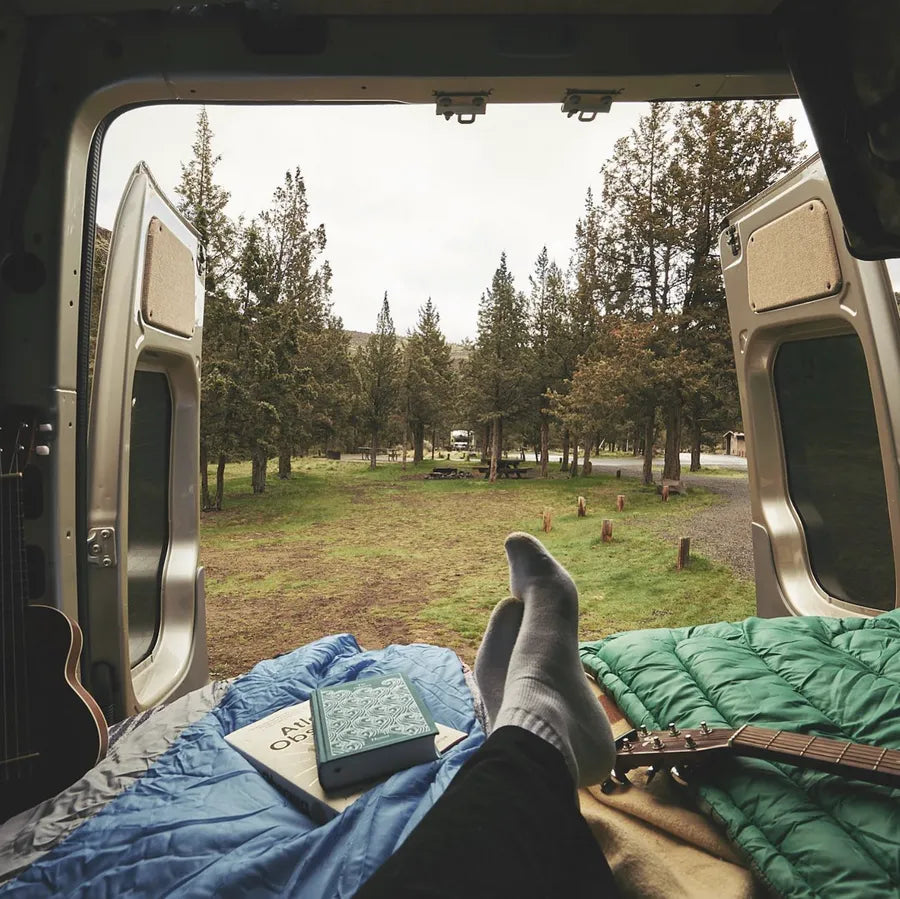 View from an Adventure Wagon camper van bed looking through the open rear doors toward a wooded campground.