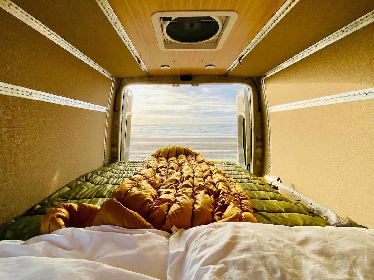Adventure Wagon camper van bed with blankets facing open rear doors and an ocean view, showing a comfortable modular van camping setup.