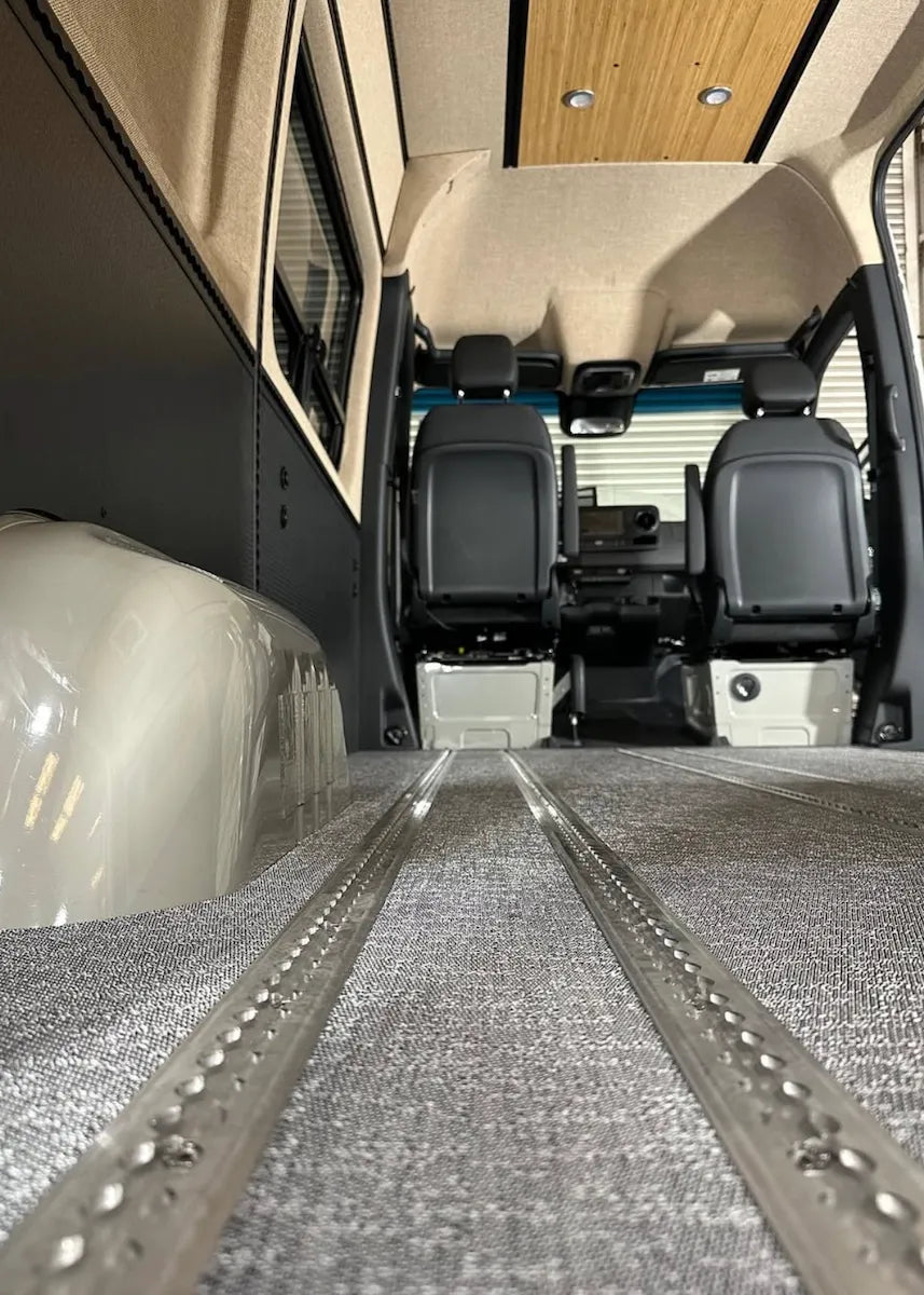 Empty Adventure Wagon camper van interior viewed from the rear, showing carpeted flooring with integrated L-track, upholstered wall panels, bamboo ceiling, and front captain’s seats.