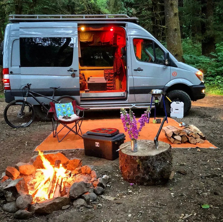 Adventure Wagon camper van parked at a forest campsite beside a campfire with camping gear set up outside.