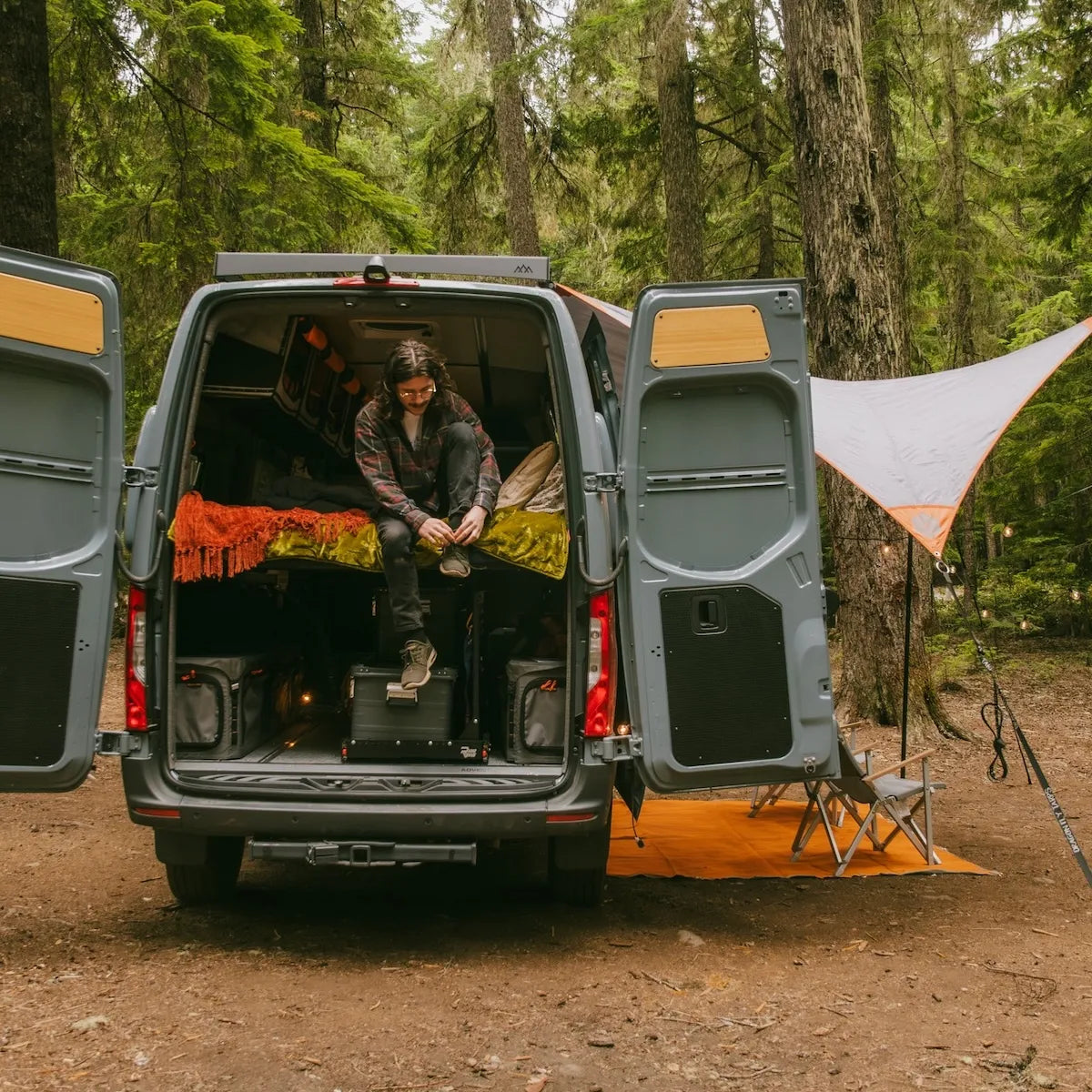 Adventure Wagon camper van parked at a forest campsite with the rear doors open, sleeping area set up, and outdoor camp shelter beside the van.