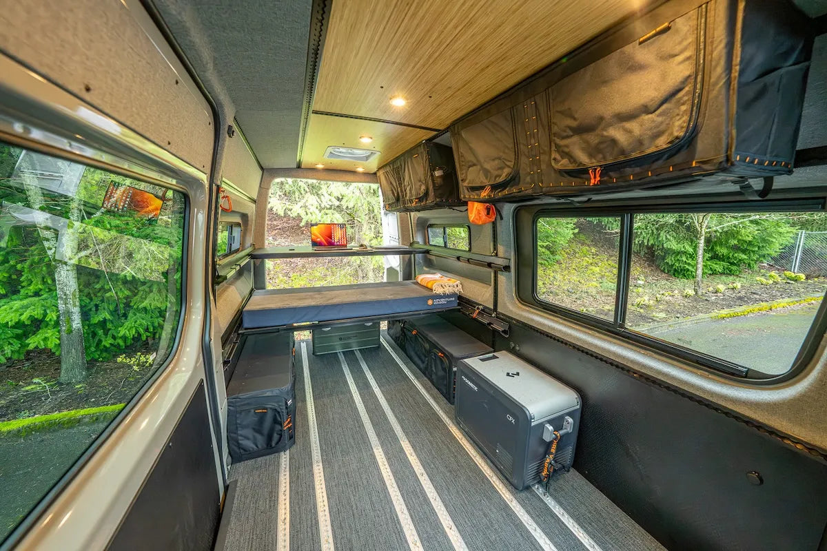 Adventure Wagon camper van interior with a raised sleeping platform, modular storage boxes, fridge, and flexible open floor space beneath the bed.