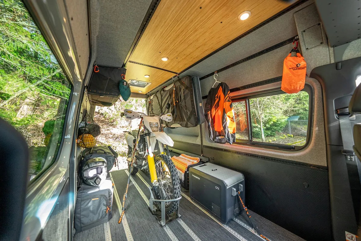Adventure Wagon camper van interior configured to transport a dirt bike with L-Track tie-downs, fridge, gear storage, and open cargo space.