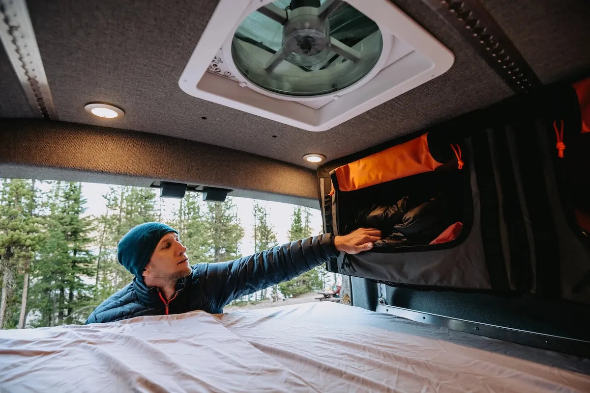 Traveler reaching into an Adventure Wagon Mule Bag that has been mounted overhead inside a camper van, providing soft-sided modular storage above the bed and living area.