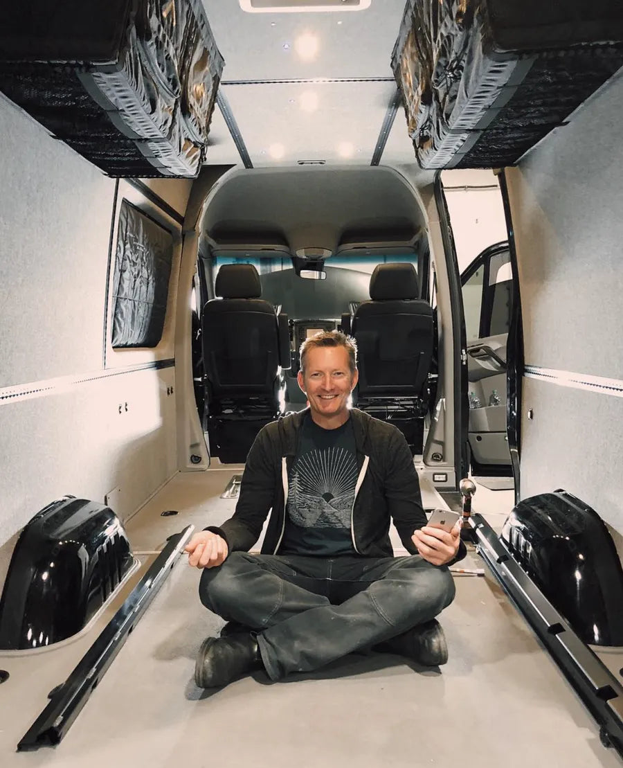 Adventure Wagon camper van owner sitting inside an empty modular interior with L-track flooring and upholstered wall panels.