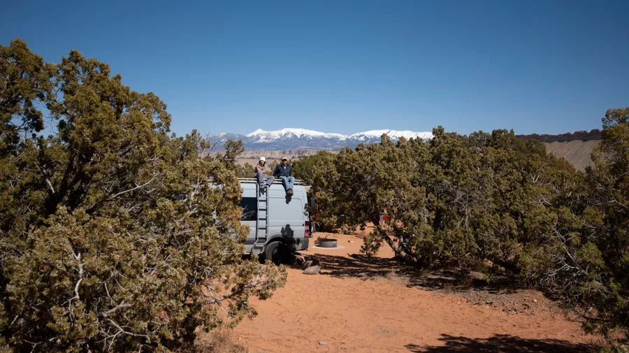 Adventure Wagon camper van parked at a mountain campsite with travelers sitting on the rooftop among desert trees.