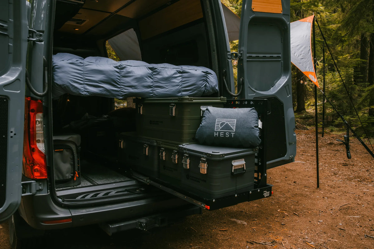 Rear view of an Adventure Wagon camper van at a forest campsite with a slide-out cargo tray loaded with Adventure Wagon storage boxes beneath the elevated bed platform.