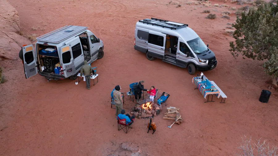 Adventure Wagon camper van at a desert campsite with campfire, outdoor seating and surrounding red rock landscape.