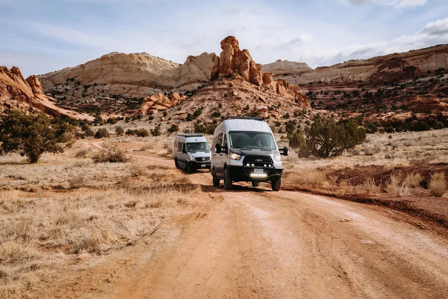 Two Adventure Wagon camper vans traveling together on a dirt road through a red rock desert landscape.