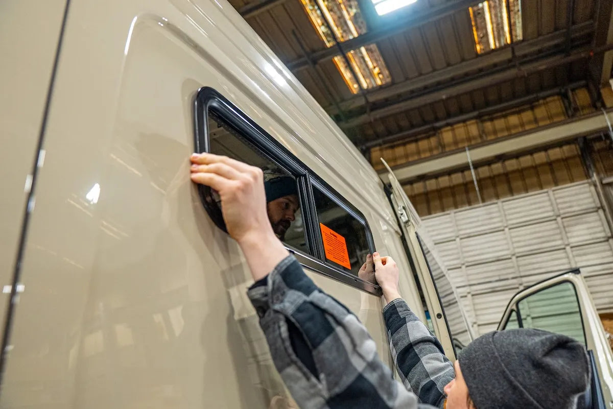 Adventure Wagon factory install technician fitting and aligning a sliding camper van window into the side of a Mercedes-Benz Sprinter.