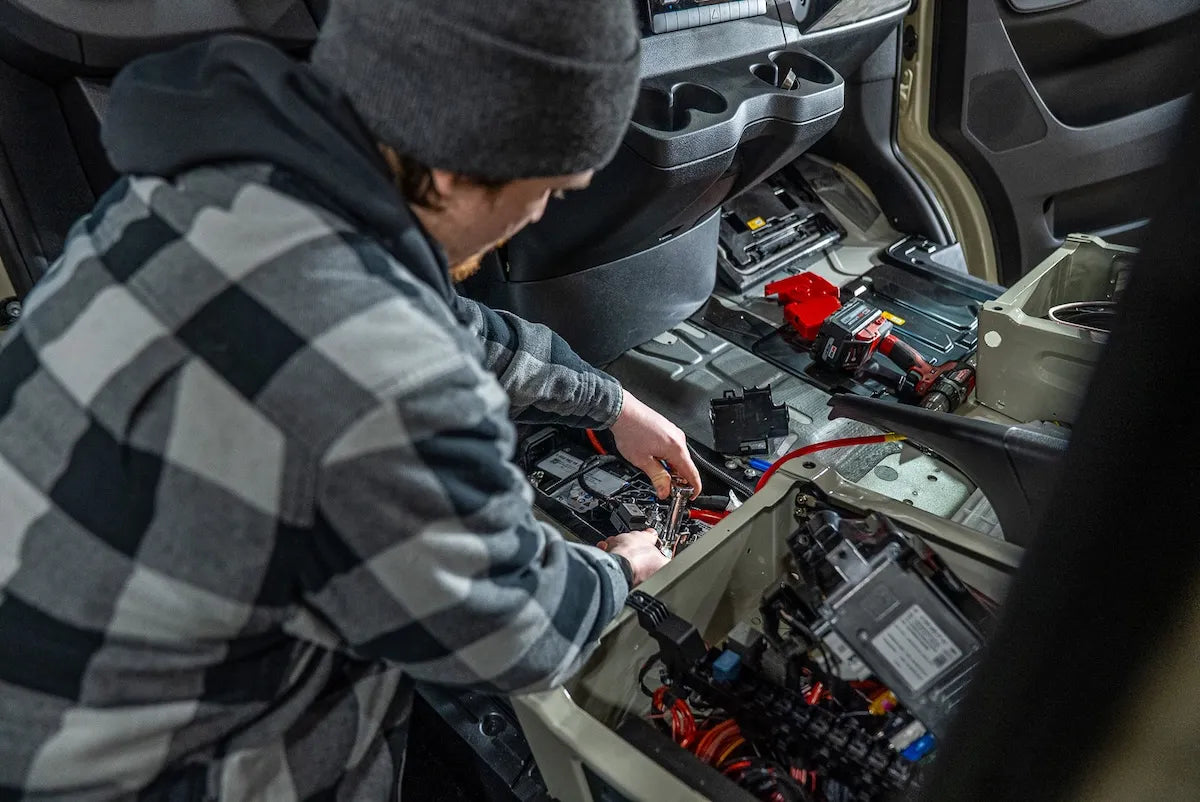 Adventure Wagon installer working on electrical wiring and components beneath the front cabin area of a camper van during Factory Install.