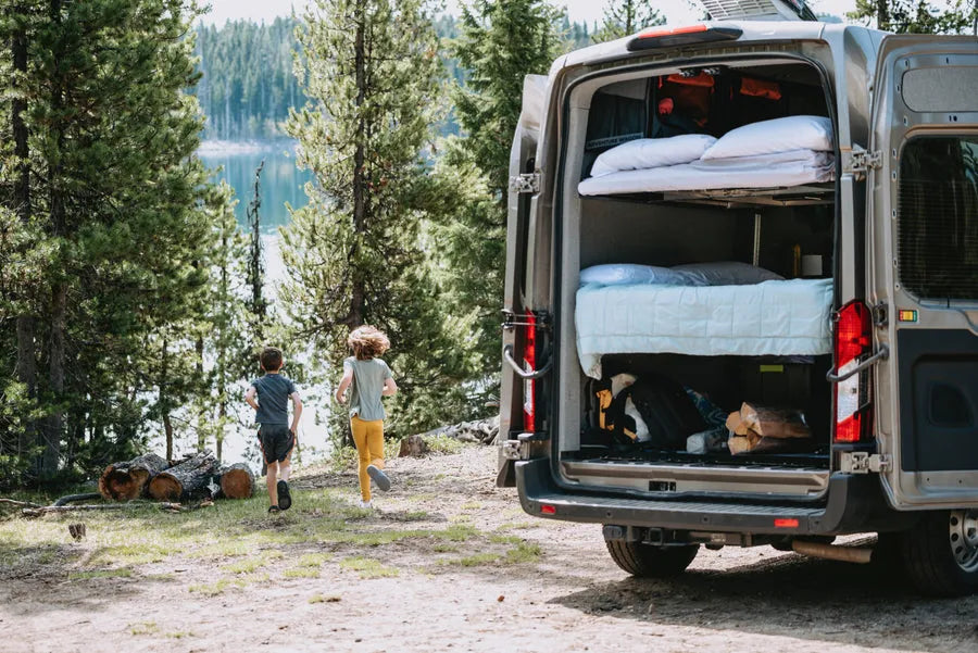 Family Adventure Wagon camper van parked at a wooded lake campsite with rear doors open and bunk beds visible inside.
