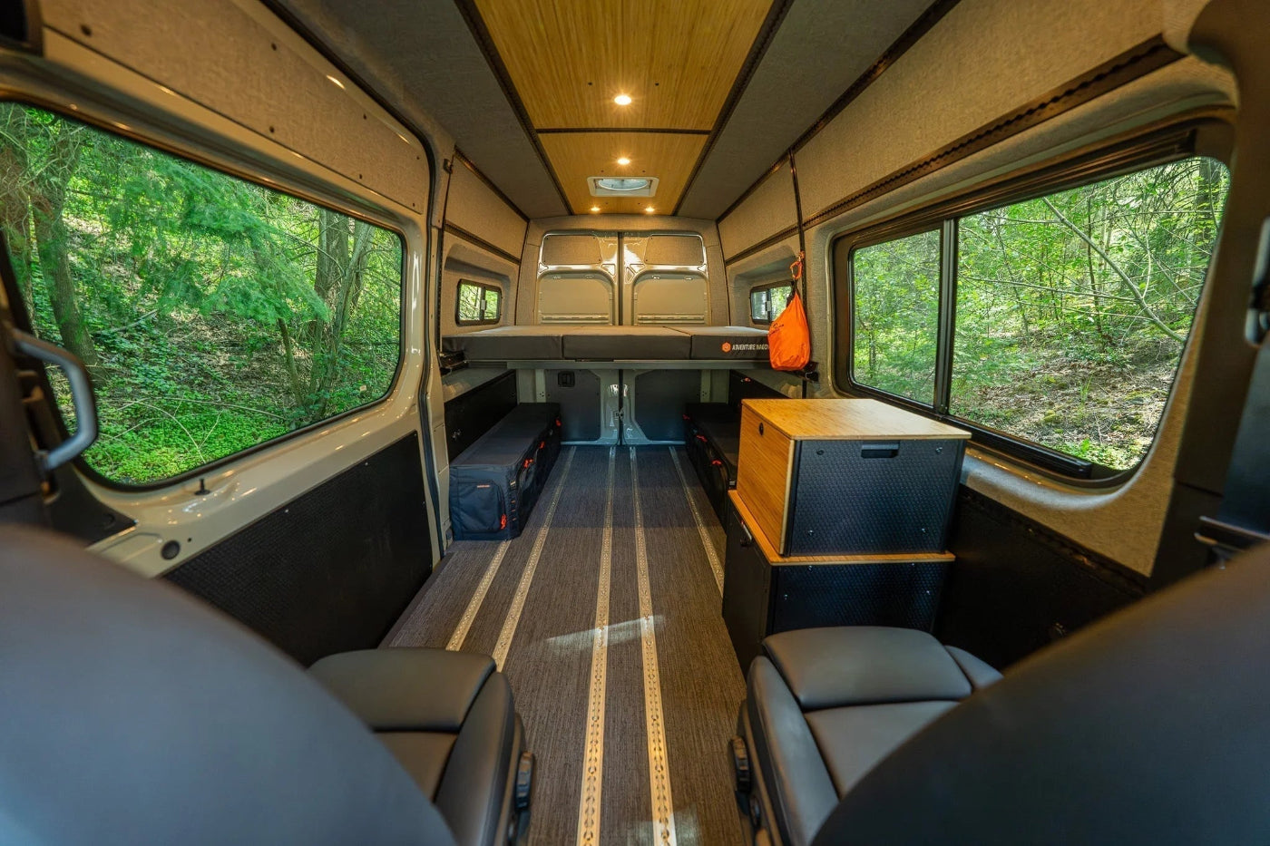 Adventure Wagon Guided Build Core RUV interior featuring a raised bed platform, modular storage, integrated L-Track flooring, and large windows overlooking the forest.
