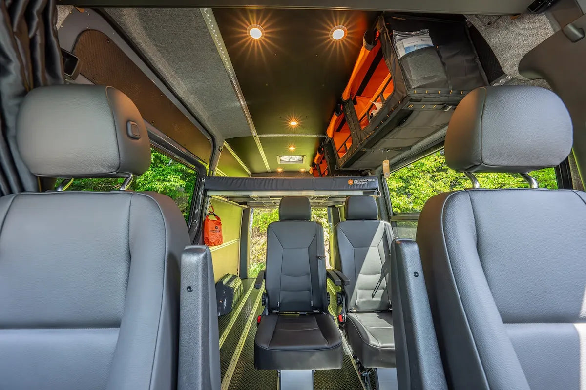 Adventure Wagon camper van interior with passenger seats, a raised rear bed platform, large windows and modular wall and ceiling panels.