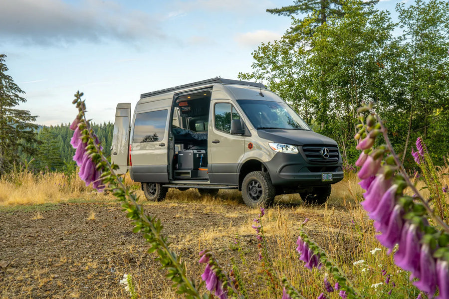 Adventure Wagon Mercedes-Benz Sprinter camper van RUV parked outdoors in a grassy mountain setting with purple wildflowers and the sliding door open.