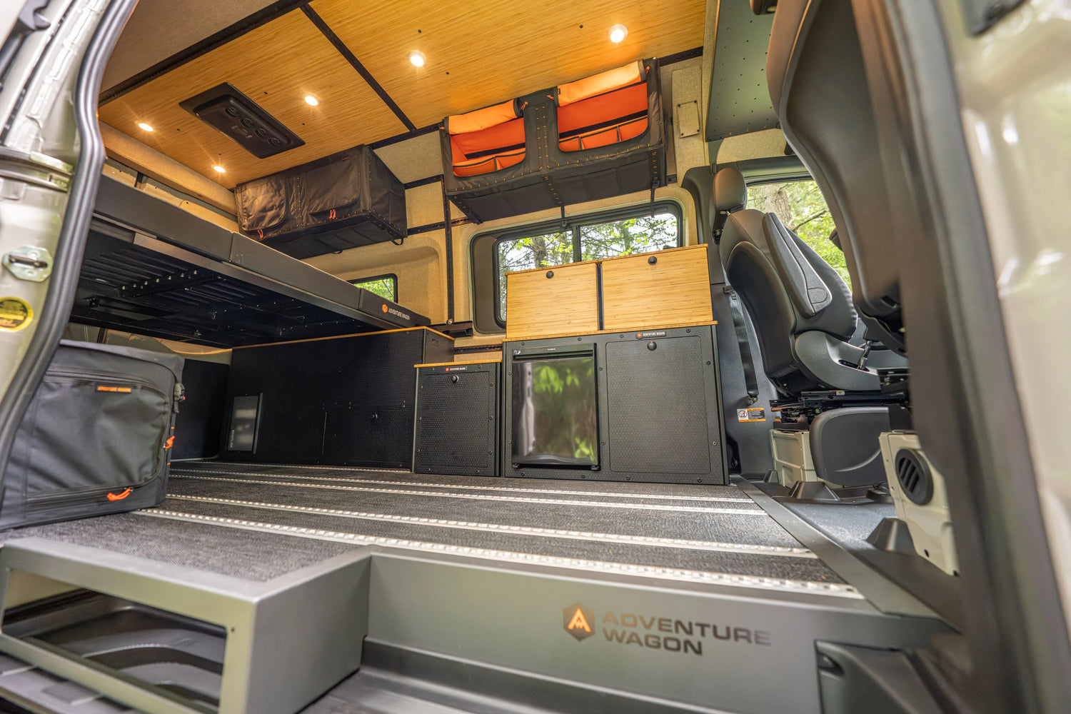 Adventure Wagon RUV 538 Mercedes-Benz Sprinter camper van interior showing the raised bed platform, Switchback Galley with bamboo cabinets and refrigerator, overhead Mule Bags, modular storage, L-track flooring, and front passenger seating.
