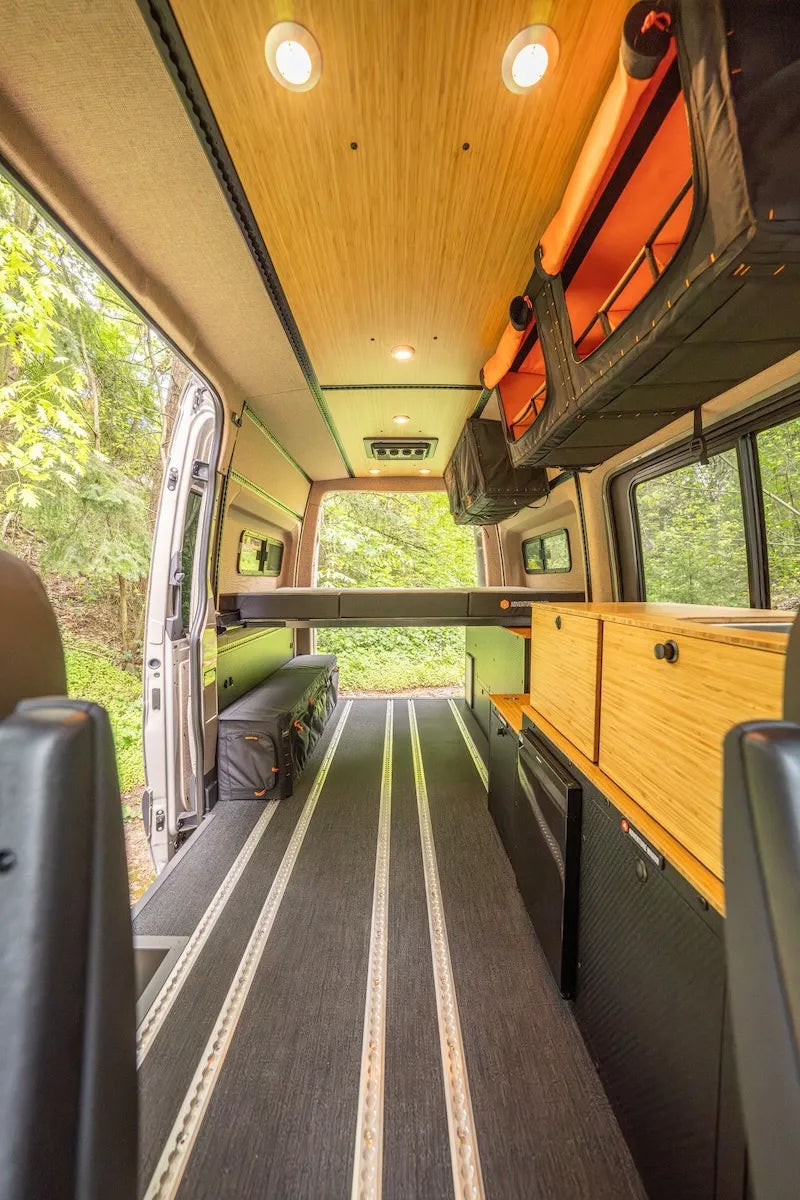 Adventure Wagon RUV camper van interior with raised bed platform, overhead Mule Bags, bamboo galley cabinetry, L-track flooring, windows, and open rear doors facing the forest.