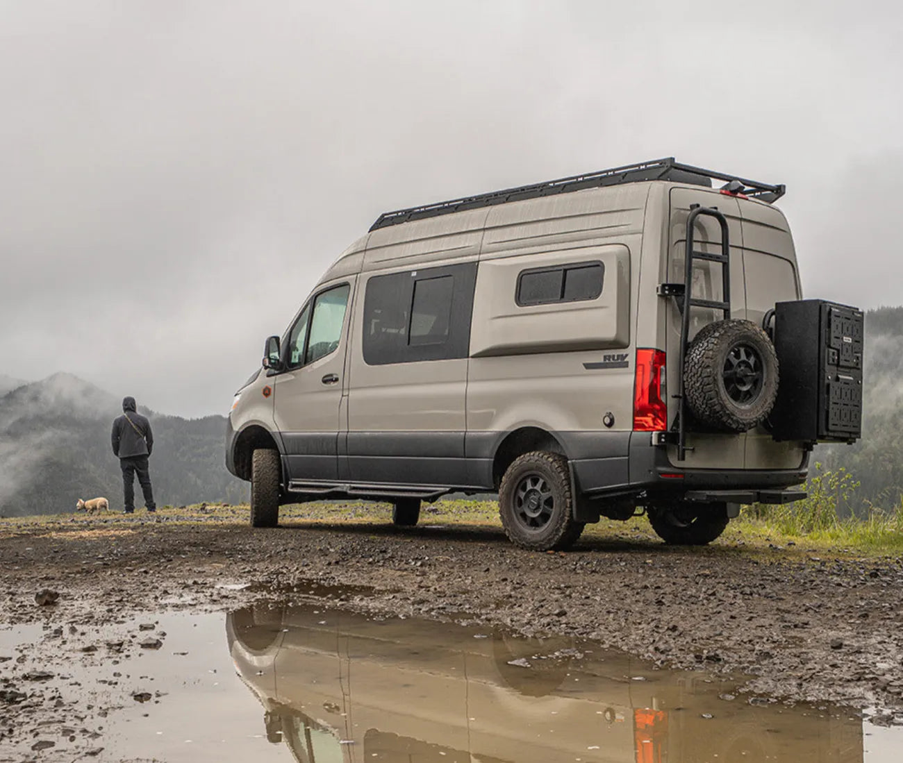 Adventure Wagon RUV camper van parked at a misty mountain overlook with off-road tires, roof rack, rear ladder, and exterior storage.