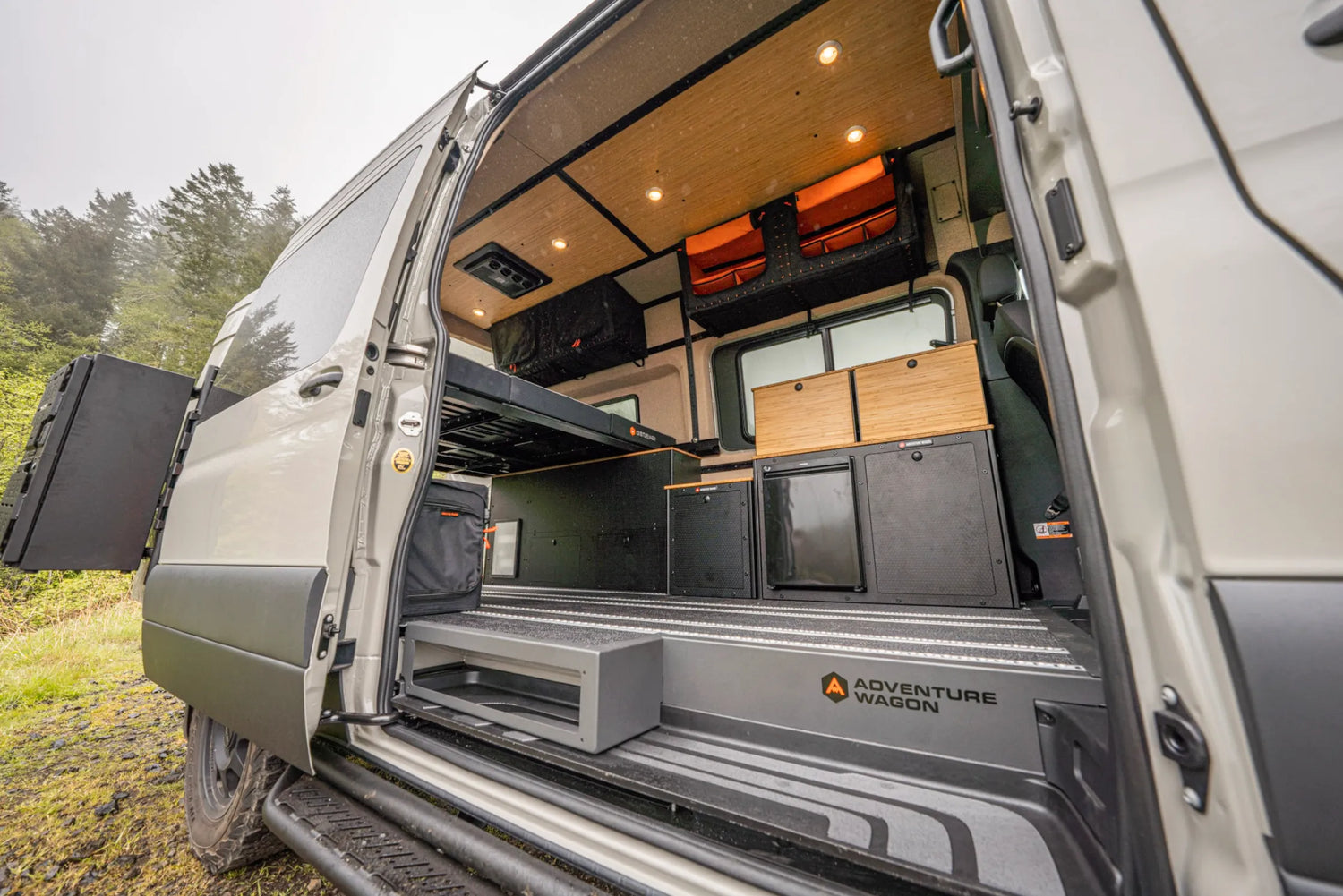 Adventure Wagon RUV Mercedes-Benz Sprinter camper van interior viewed through the open side door, showing a raised bed platform, Switchback Galley with bamboo cabinets and refrigerator, overhead Mule Bags, modular storage, L-track flooring, and bamboo ceiling panels.