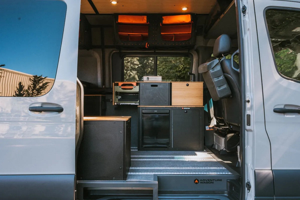 Adventure Wagon RUV camper van viewed through the open sliding side door, showing the Switchback Galley, refrigerator, overhead storage, passenger seat, and L-track flooring.
