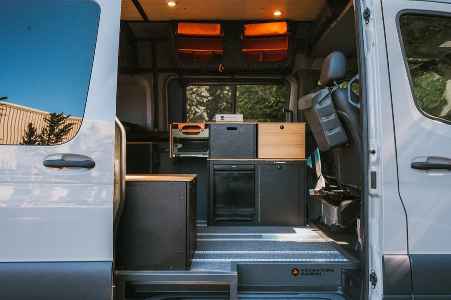 Adventure Wagon RUV camper van viewed through the open sliding side door, showing the Switchback Galley, refrigerator, overhead storage, passenger seat, and L-track flooring.