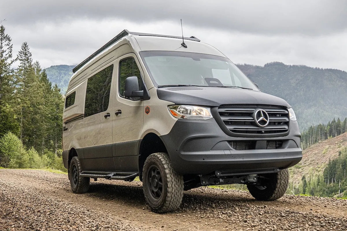 Adventure Wagon RUV Mercedes-Benz Sprinter camper van with off-road tires parked on a gravel mountain road overlooking forested hills.