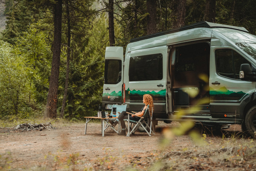 Solo traveler relaxing beside the Adventure Wagon Solo Nomad camper van at a forest campsite.