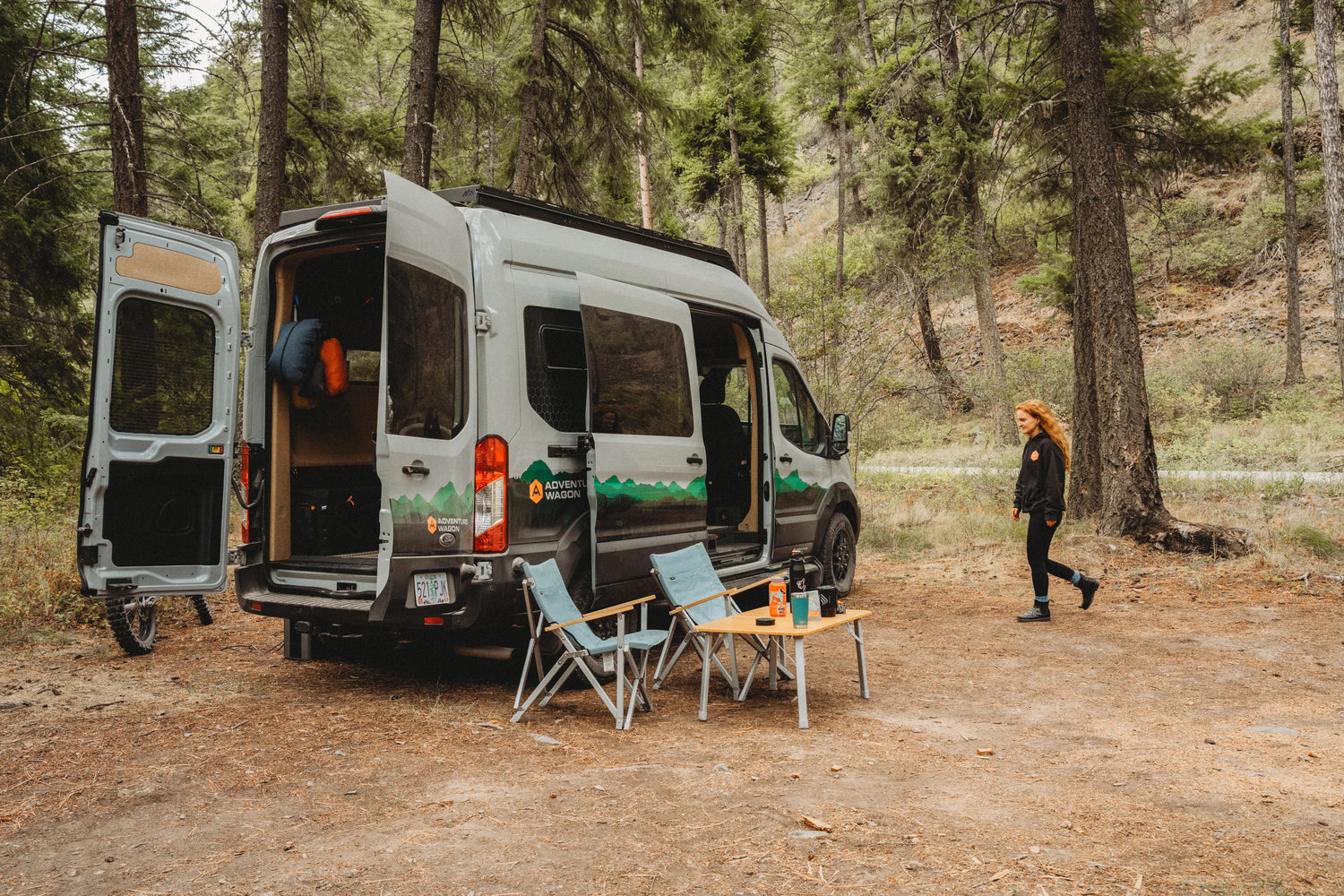 Adventure Wagon Solo Nomad camper van set up at a forest campsite with chairs and an outdoor table.