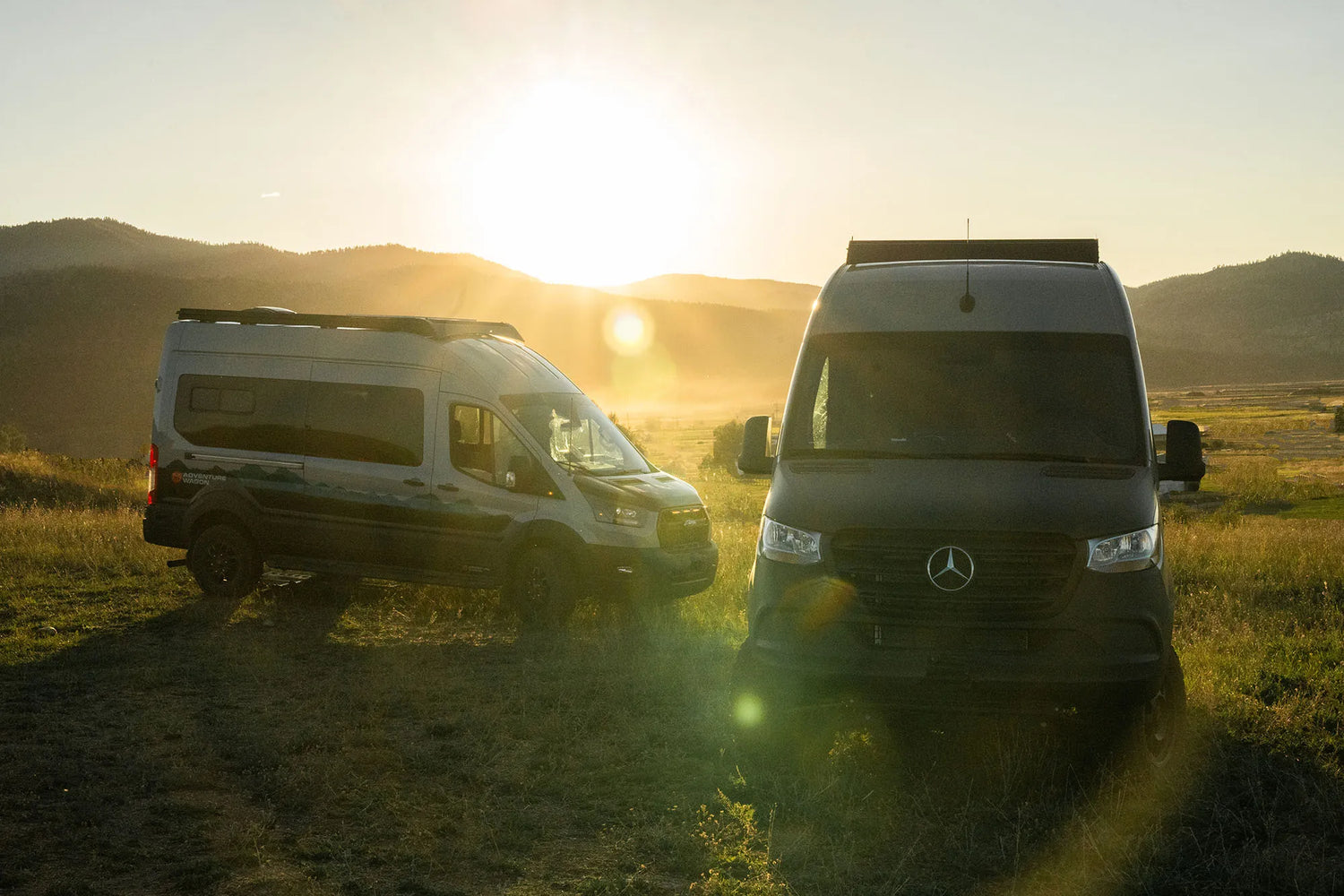 Two camper vans, specifically sprinter and transit vans, parked in a field with a sunset in the background