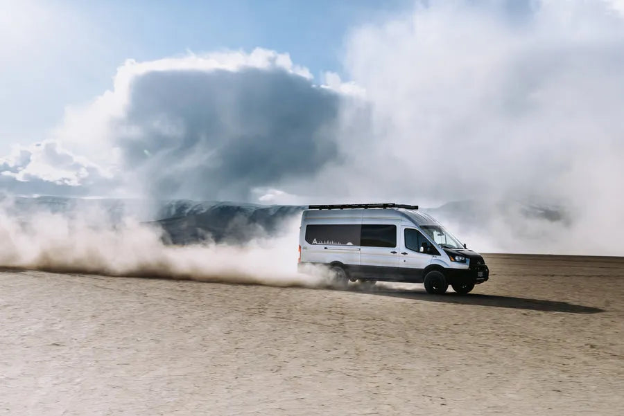 Adventure Wagon Mercedes Sprinter camper van driving across a dusty off-road landscape with a large dust cloud behind it.