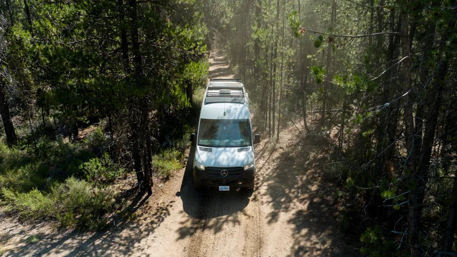 Adventure Wagon Mercedes Sprinter camper van traveling along a forest road with rear-mounted spare tire and recovery gear.
