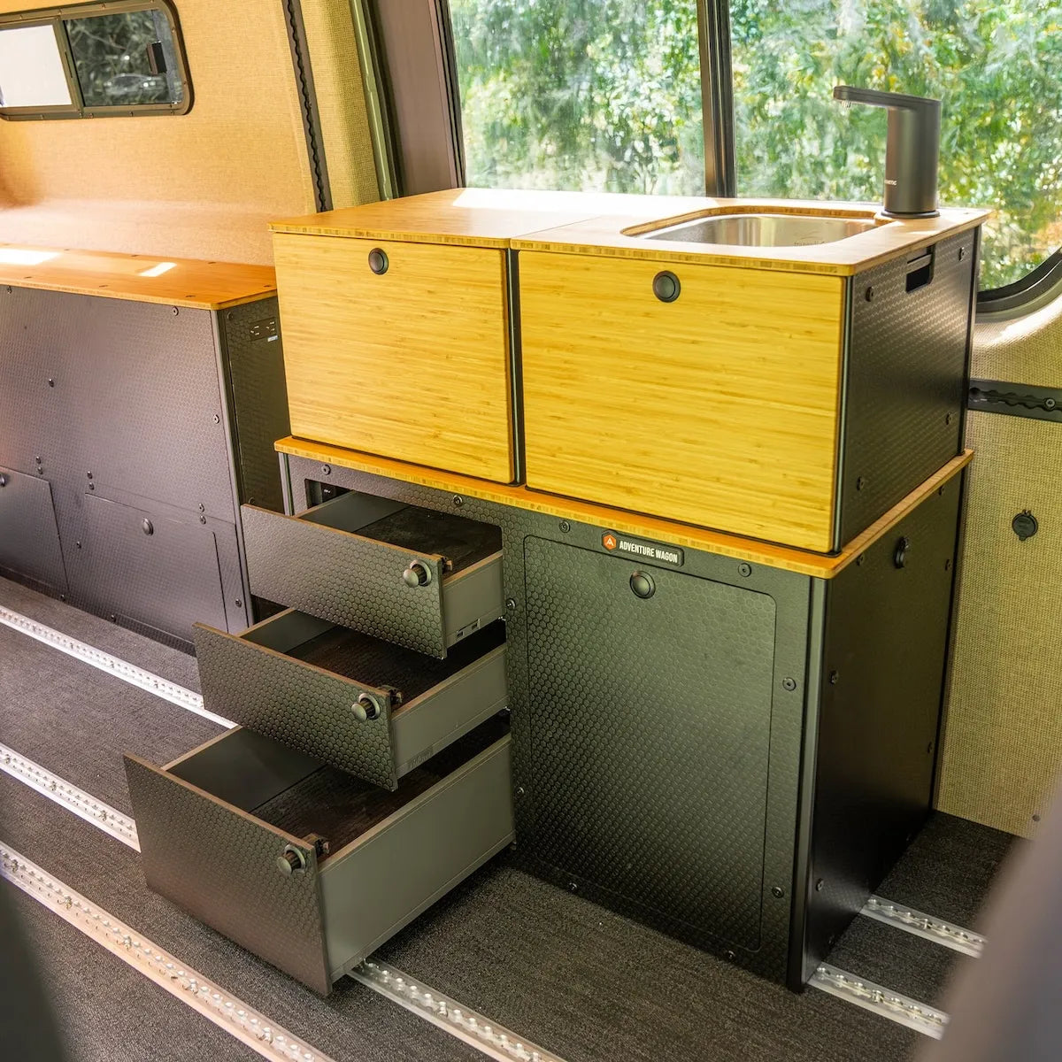 Adventure Wagon Switchback Galley camper van kitchen with bamboo cabinet fronts, stainless sink, three open storage drawers, and modular black cabinetry.