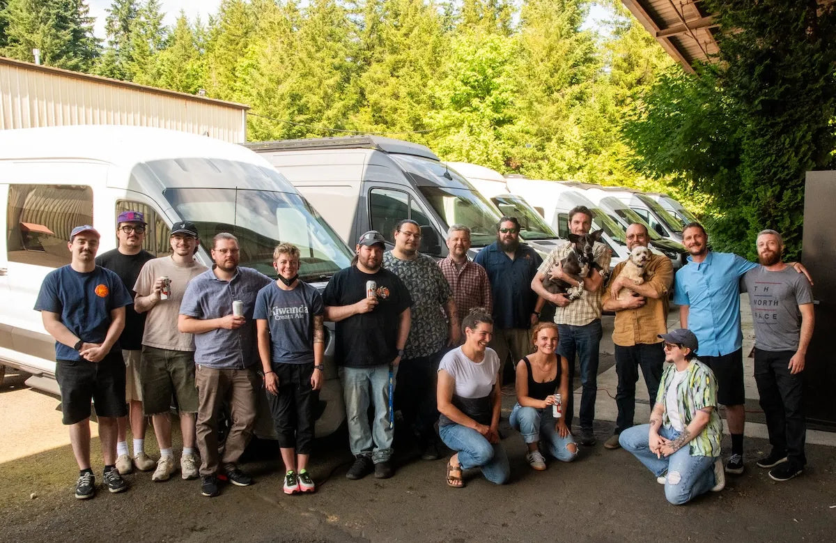 Adventure Wagon team members posing together in front of a lineup of Mercedes-Benz Sprinter camper vans outside the shop.