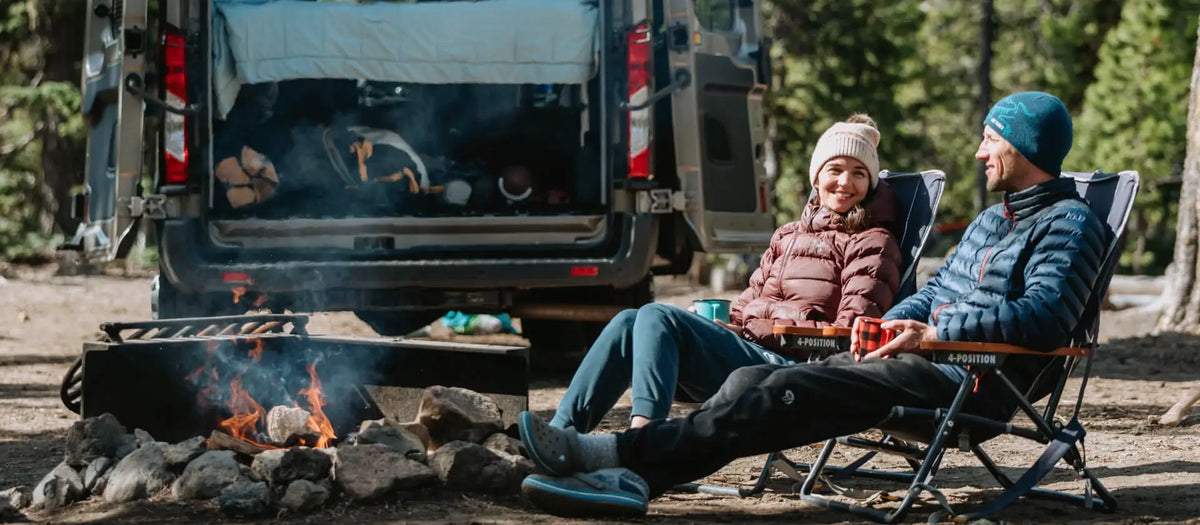 Adventure Wagon. A couple relaxes in folding chairs by a campfire at a forest campsite, with an open camper van behind them. The person on the left wears a pink puffer jacket and beige beanie, and the person on the right wears a blue puffer jacket and dark beanie.