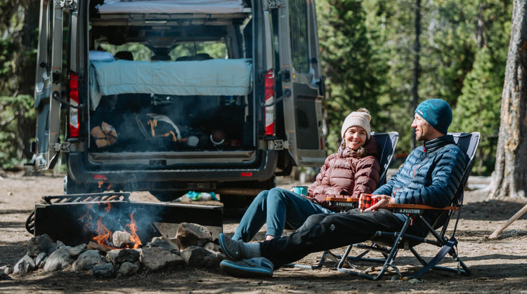 Two people sitting in outdoor chairs around a campfire with their Adventure Wagon modular van behind them 